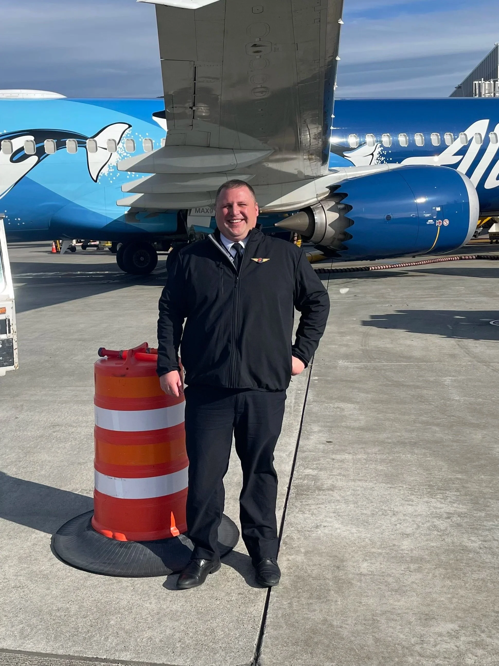 A smiling man in a pilot uniform standing on an airport tarmac near an airplane painted with a dolphin and ocean waves design. He is next to an orange and white striped safety cone and surrounded by ground equipment.