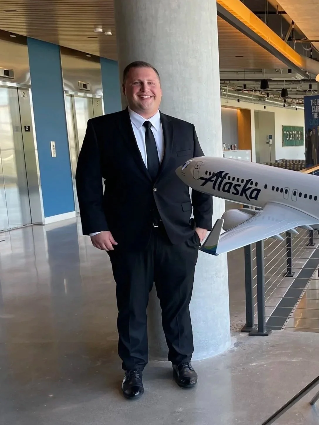 A man in a black suit and tie standing inside an airport next to a model airplane labeled 'Alaska Airlines.' He is smiling and has one hand in his pocket, with elevators and modern interior decor visible in the background.