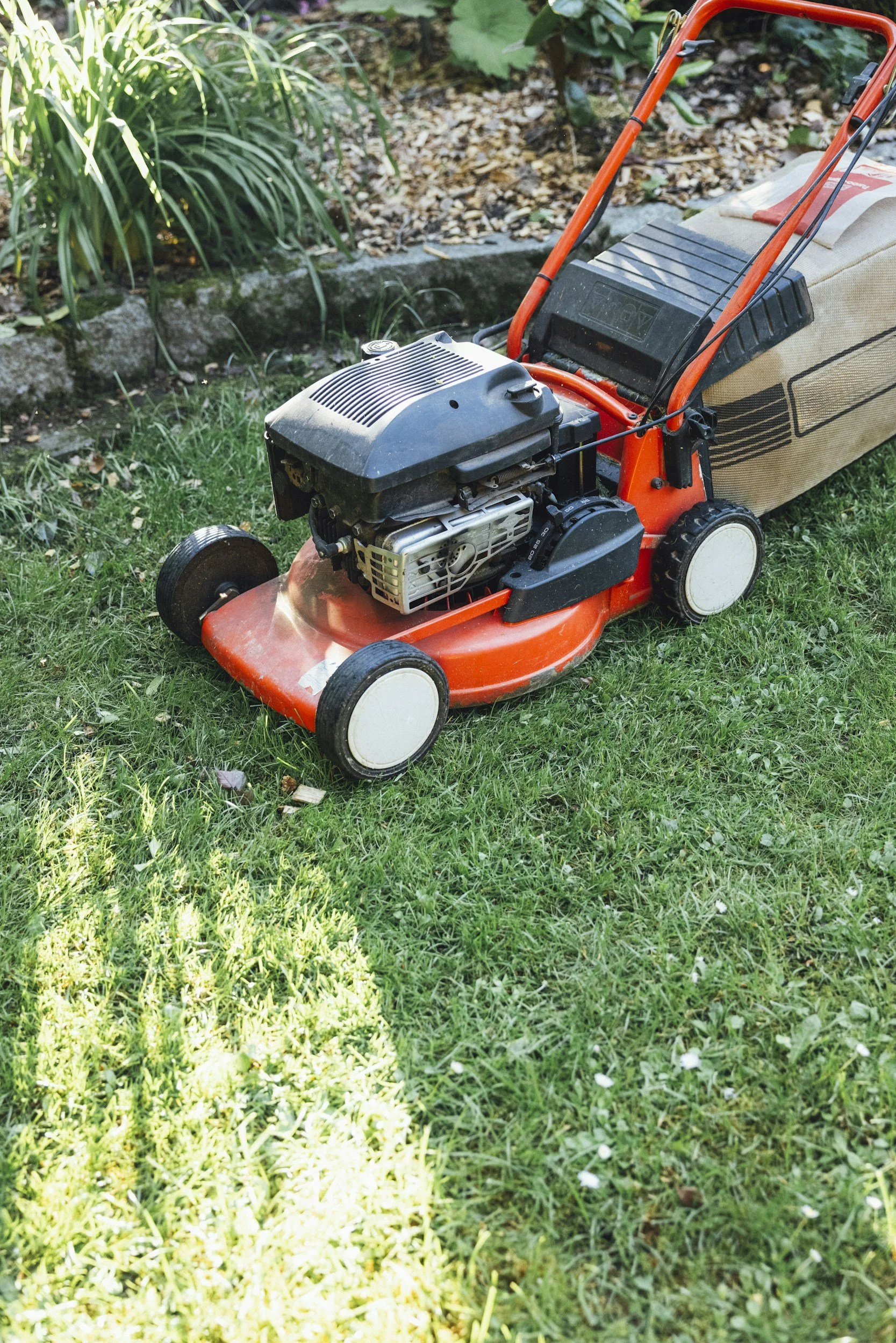 A lawnmower with a red handle and black engine is sitting on green grass in a garden, next to a flower bed with bushes and fallen leaves.