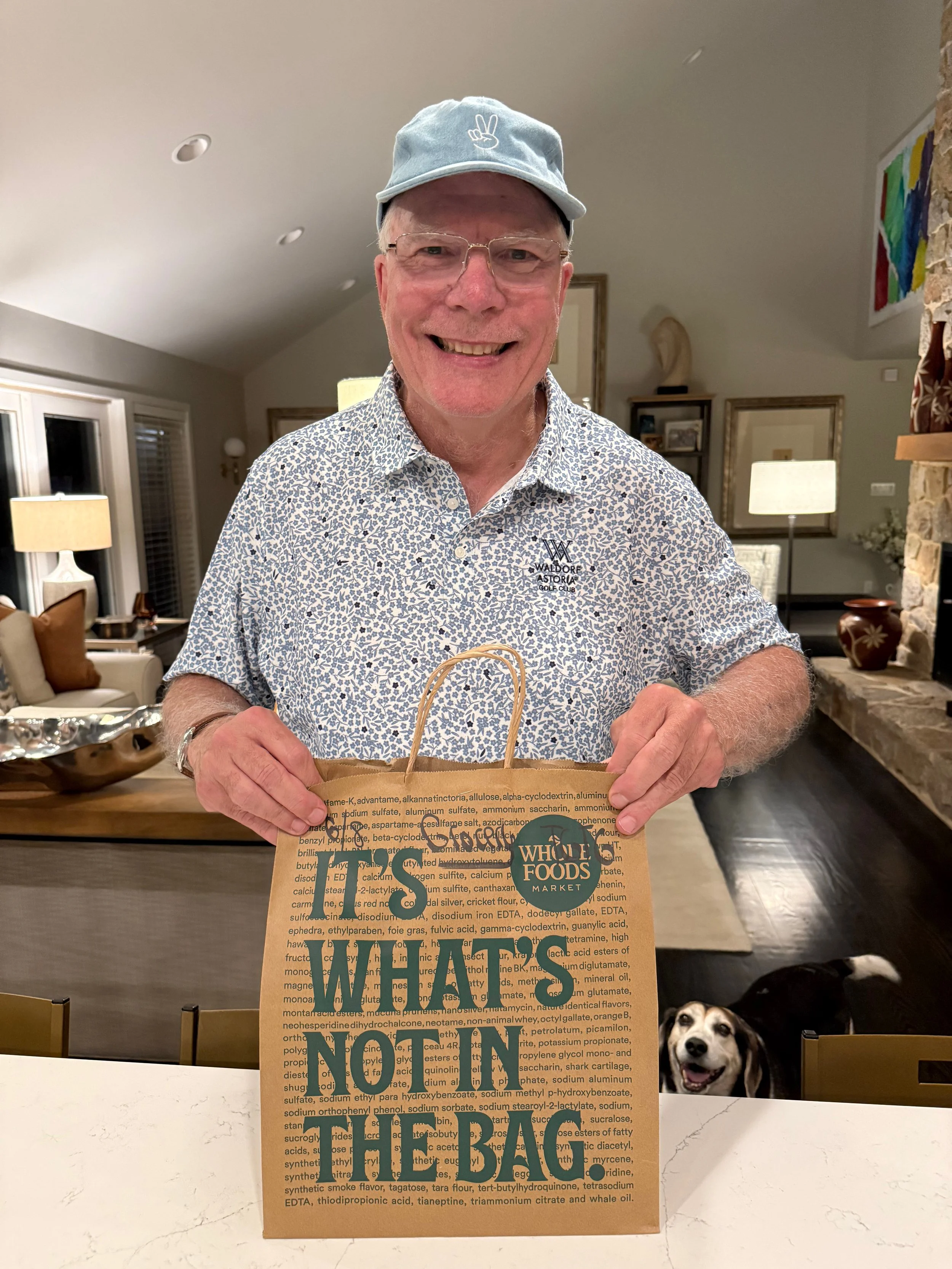 A smiling man in a floral shirt and glasses is holding a Whole Foods Market paper bag in a well-lit living room. There are two dogs peeking over a marble counter in the foreground.