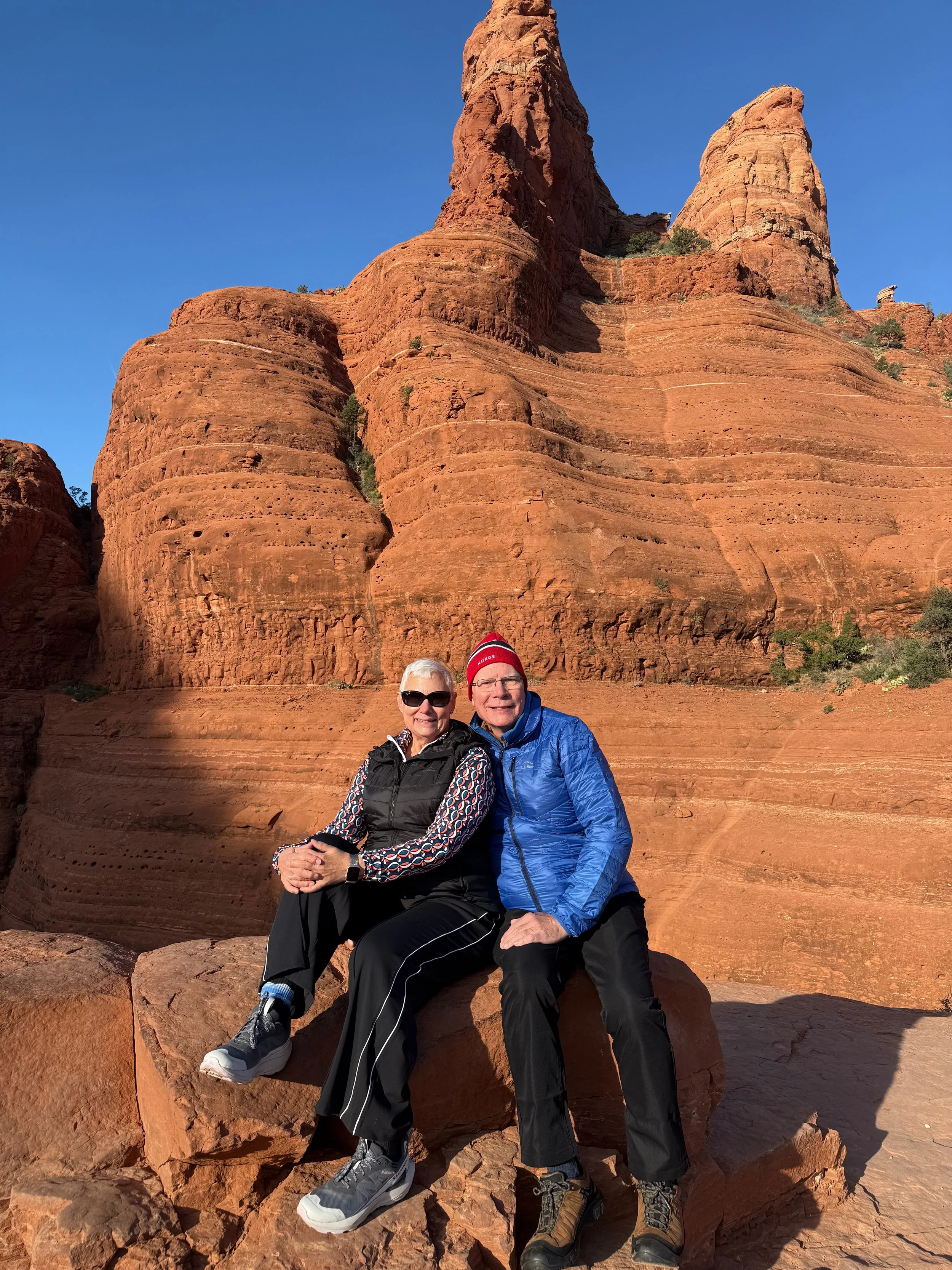 A smiling senior couple sitting on a large rock in front of tall red sandstone formations with a clear blue sky in the background.