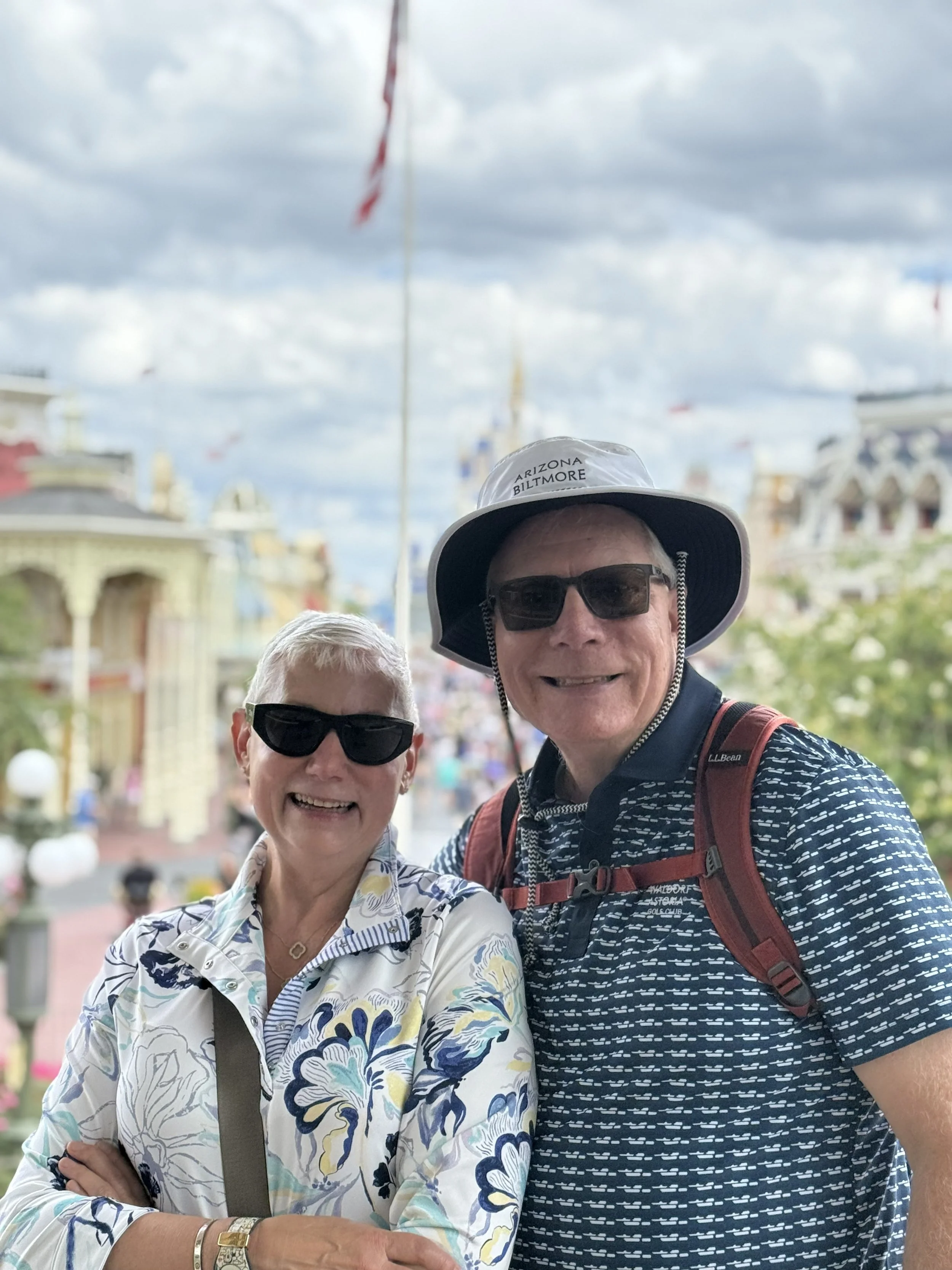 A smiling elderly woman and man pose together in an outdoor setting with cloudy skies, colorful buildings, and flags in the background. The woman has short gray hair, is wearing sunglasses, a floral blouse, and jewelry. The man has a broad smile, is wearing sunglasses, a hat with