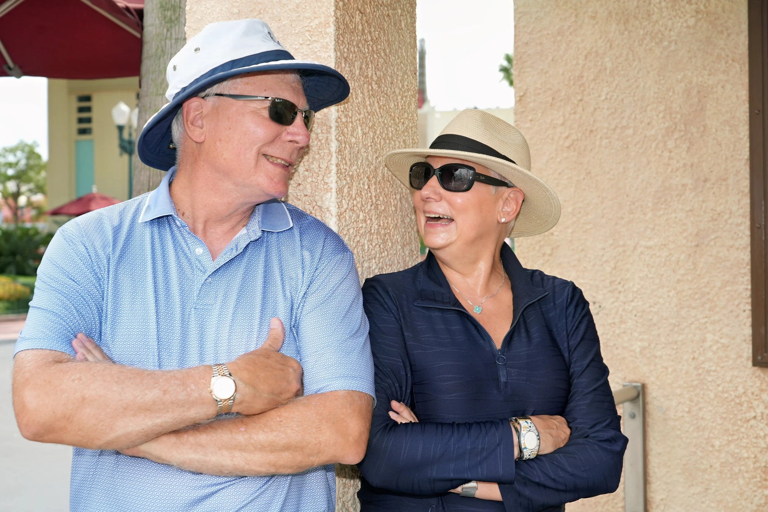An older man and woman smiling and talking, wearing hats and sunglasses, standing outdoors against a beige wall.