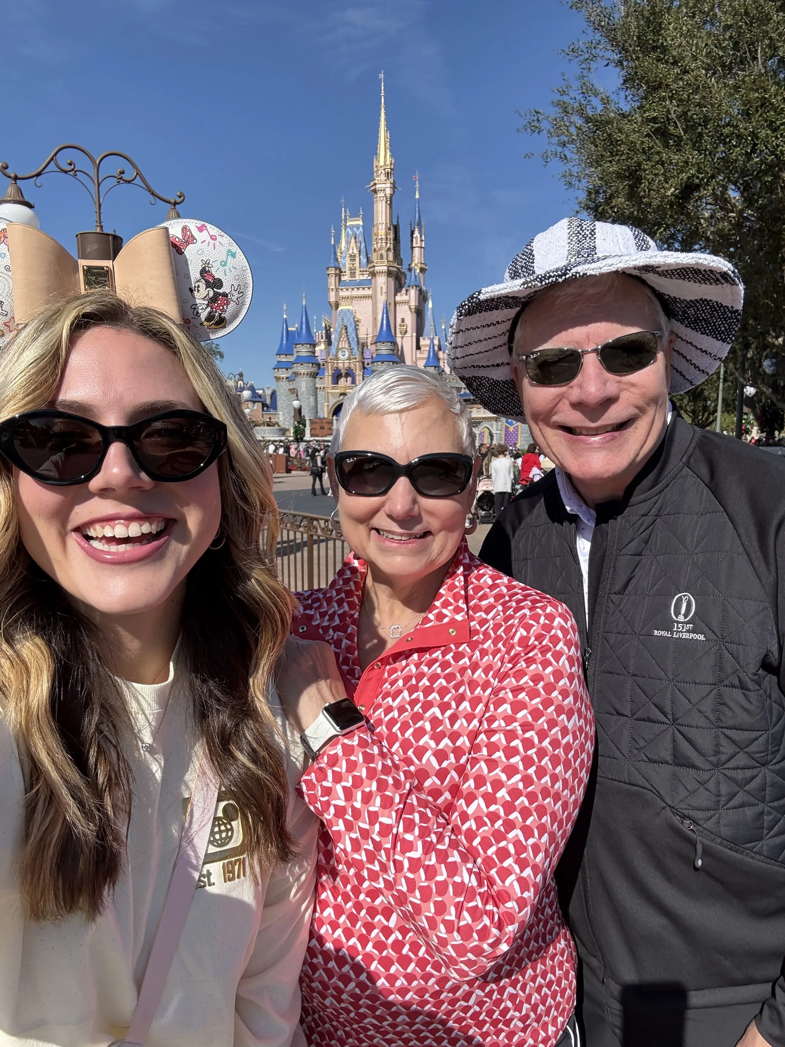 Three people smiling at Disneyland with Sleeping Beauty's Castle in the background. The woman on the left has blonde hair, wearing sunglasses, and Mickey Mouse ears hat. The woman in the middle has short gray hair, wearing glasses and a red patterned shirt. The man on the right has light hair, wearing sunglasses, a striped wide-brim hat, and a black quilted jacket.