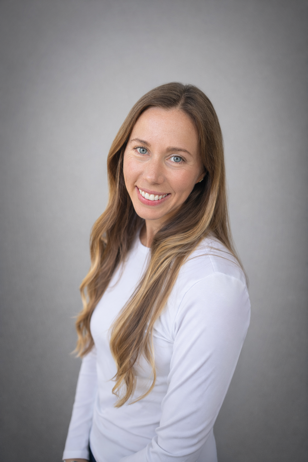 Smiling woman with long wavy blond hair, wearing a white top, against a gray background.