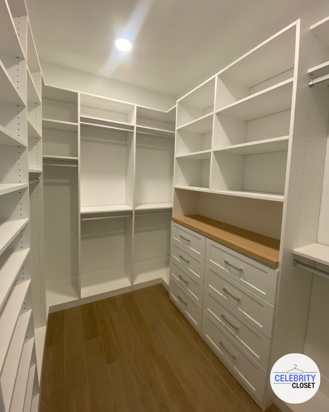 Empty walk-in closet with white shelves and drawers, wooden floor, ceiling light, and the logo 'Celebrity Closet'.