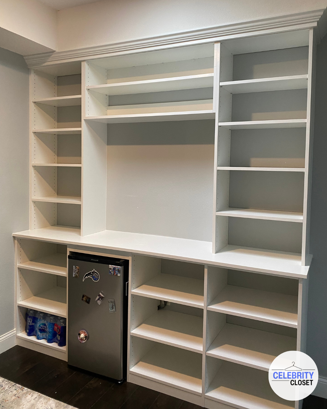 White built-in shelving unit with open shelves, a countertop, a mini fridge with a Maryland Ravens sticker and magnets, and bottled water stored on the bottom left shelf.