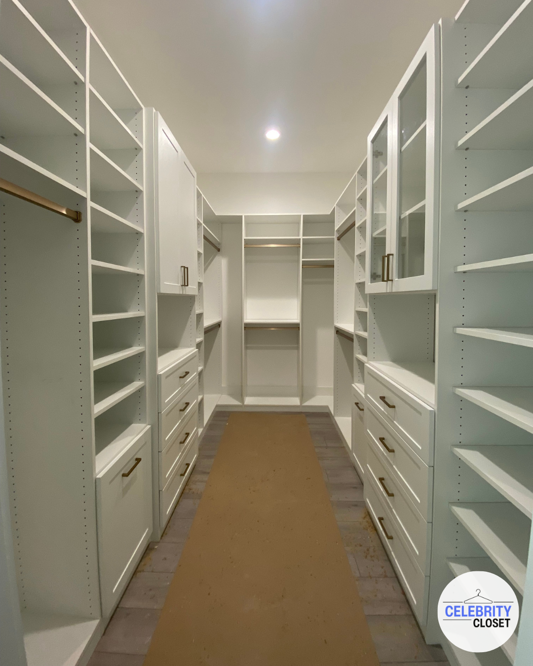 Empty walk-in closet with white shelves and drawers, gold handles, and hardwood flooring, with a brown rug in the center.