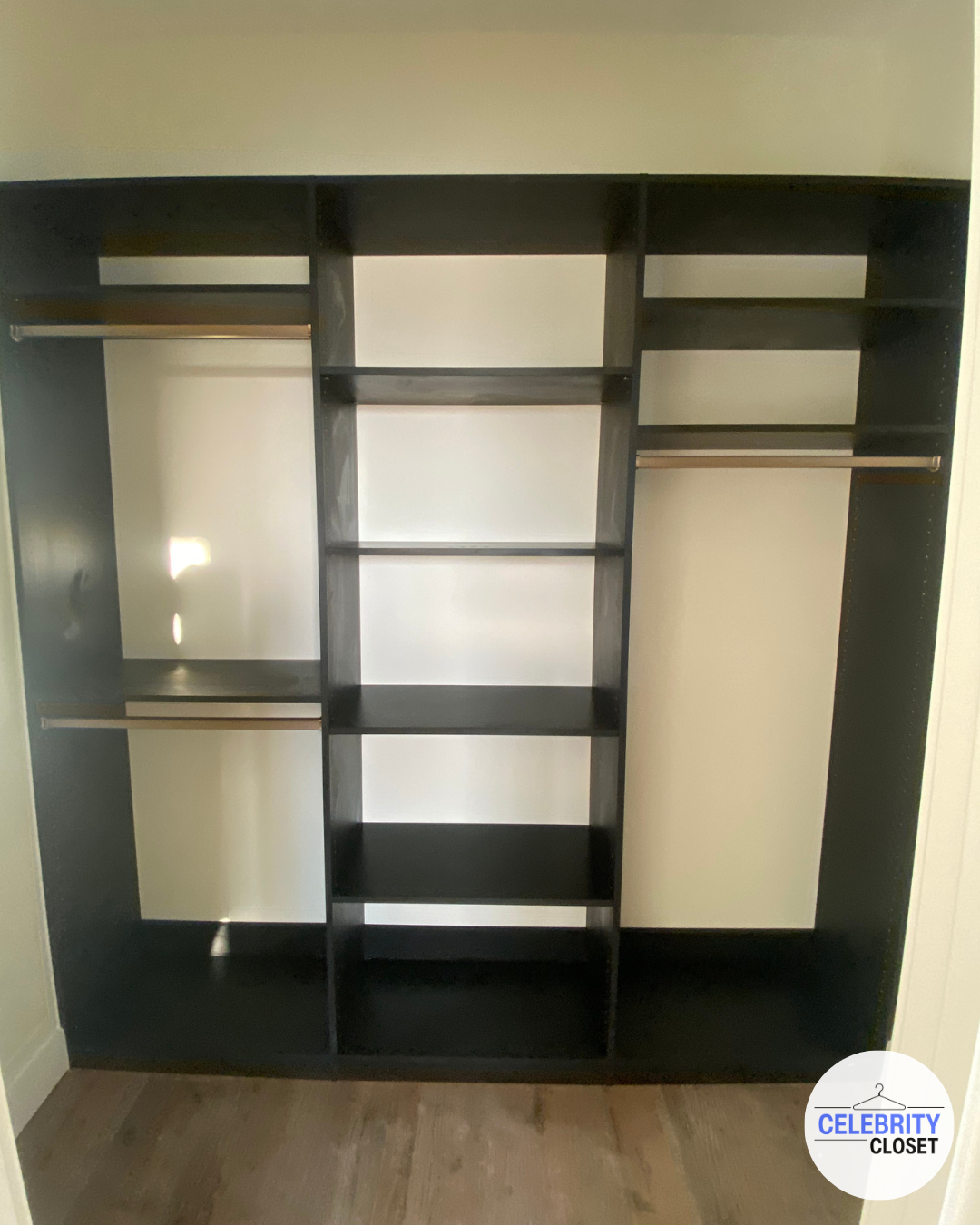 Empty black closet organizer with multiple shelves and hanging rods, placed against a white wall near a wooden floor.