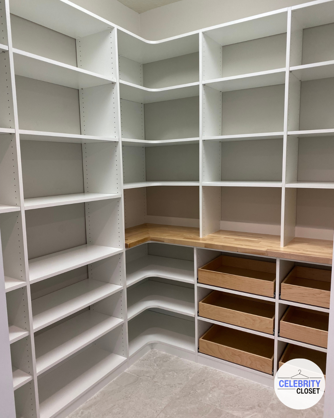 Empty white closet with numerous shelves, drawers, and a wooden countertop, in a corner of a room.