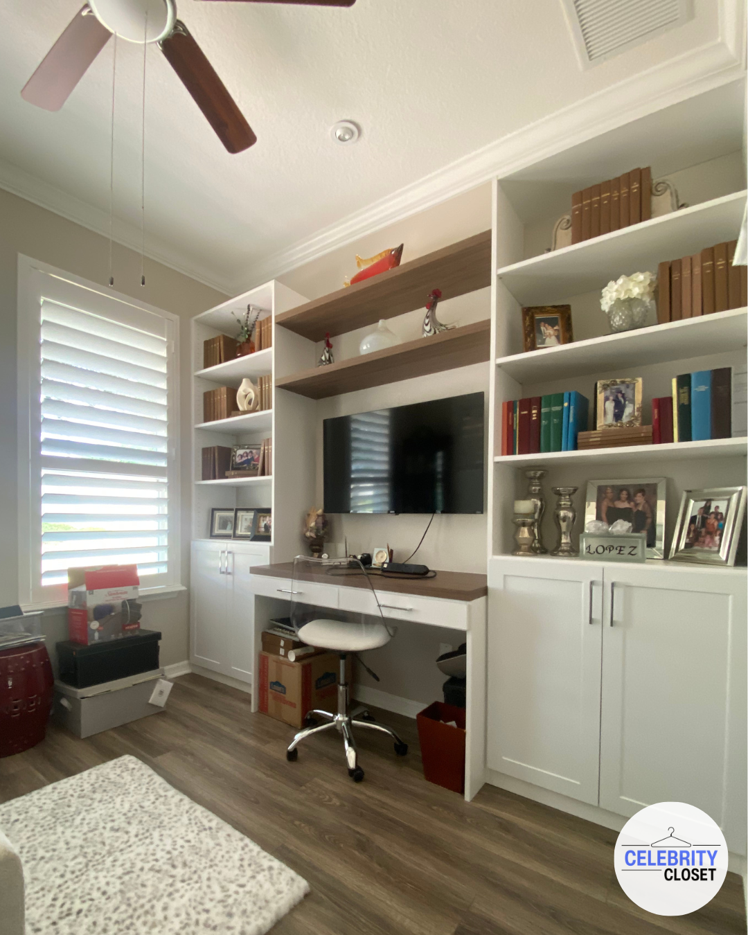 Living room with white built-in shelves and cabinets, a flat-screen TV, and decorative items like books and photo frames. A small desk with a swivel chair is positioned below the TV, with various boxes and storage containers nearby. The room has wood