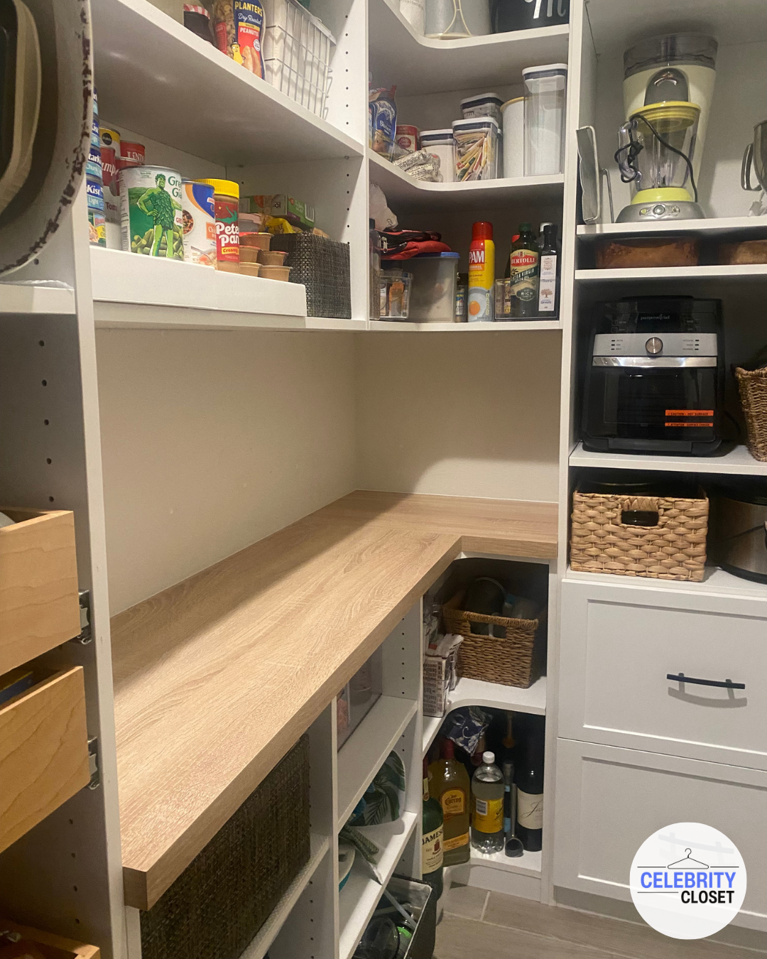 Organized pantry storage with shelves containing various canned goods, jars, bottles, baskets, kitchen appliances, and a wooden countertop.