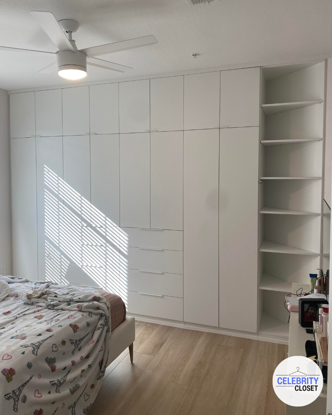 Bedroom with white closet that has multiple doors and drawers, a corner open shelf, part of a bed with a colorful Paris-themed blanket, and a ceiling fan with a light. Sunlight filters through window blinds casting striped shadows.