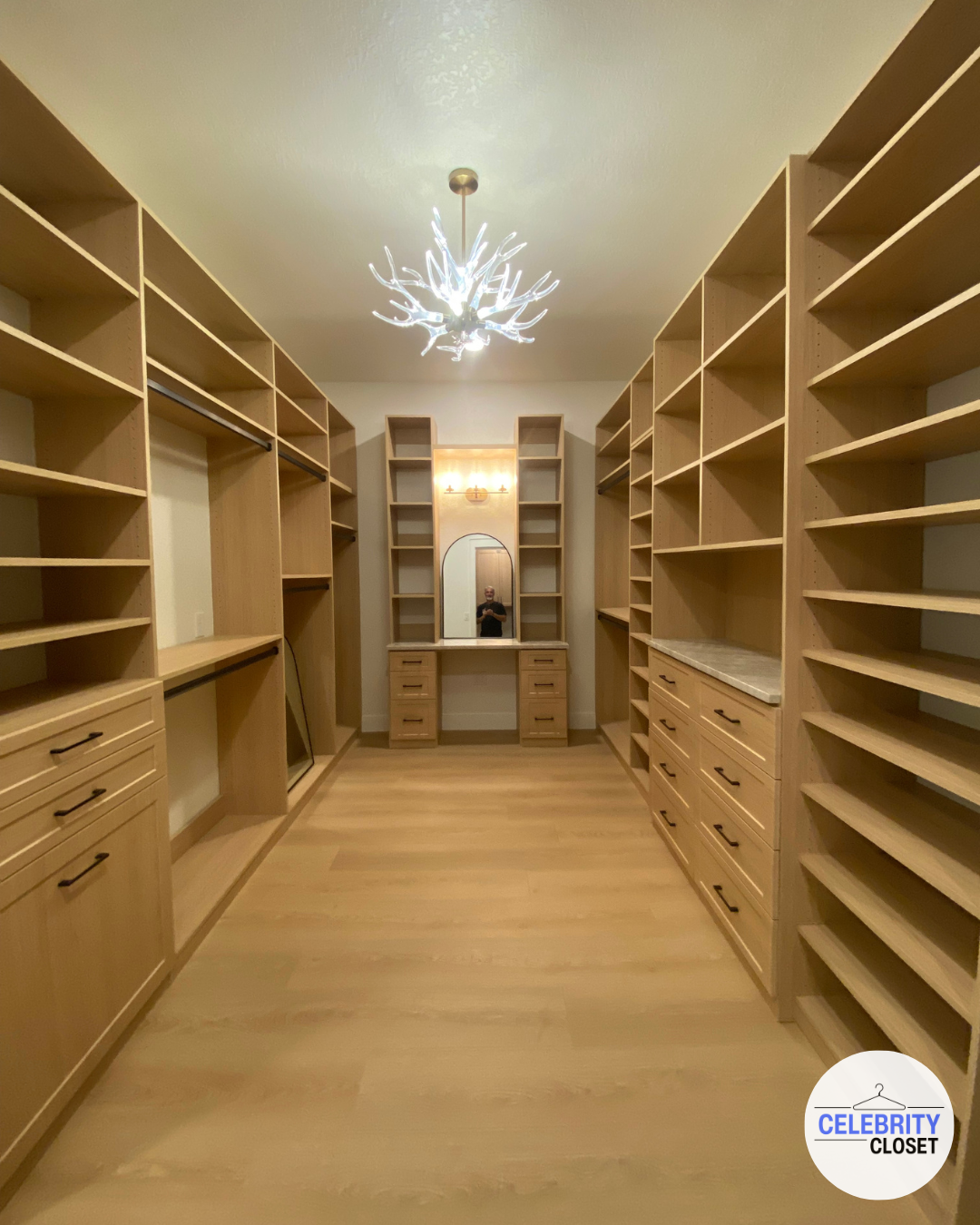 Empty walk-in closet with wooden shelves and drawers on both sides, a small vanity with a mirror and drawer units at the back, and a modern chandelier hanging from the ceiling.