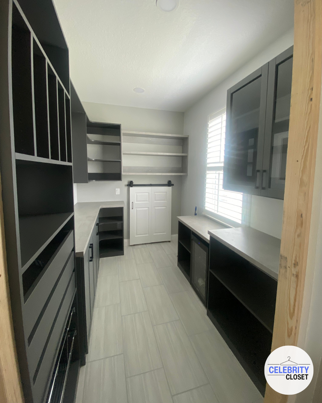 A small room with black and gray cabinets, open shelves, a countertop, and a window with white blinds. The floor has light-colored tiles and a logo in the bottom right corner says 'Celebrity Closet'.