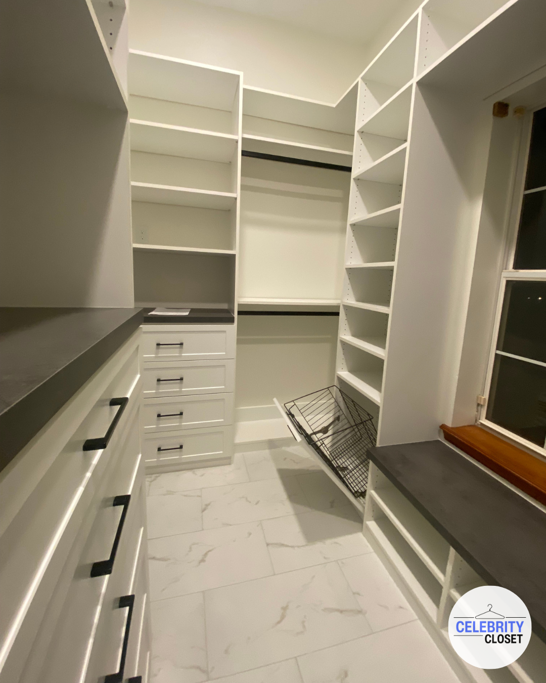 A walk-in closet with white shelving, drawers, and black handles, a countertop, and a wire basket on the floor near the window.