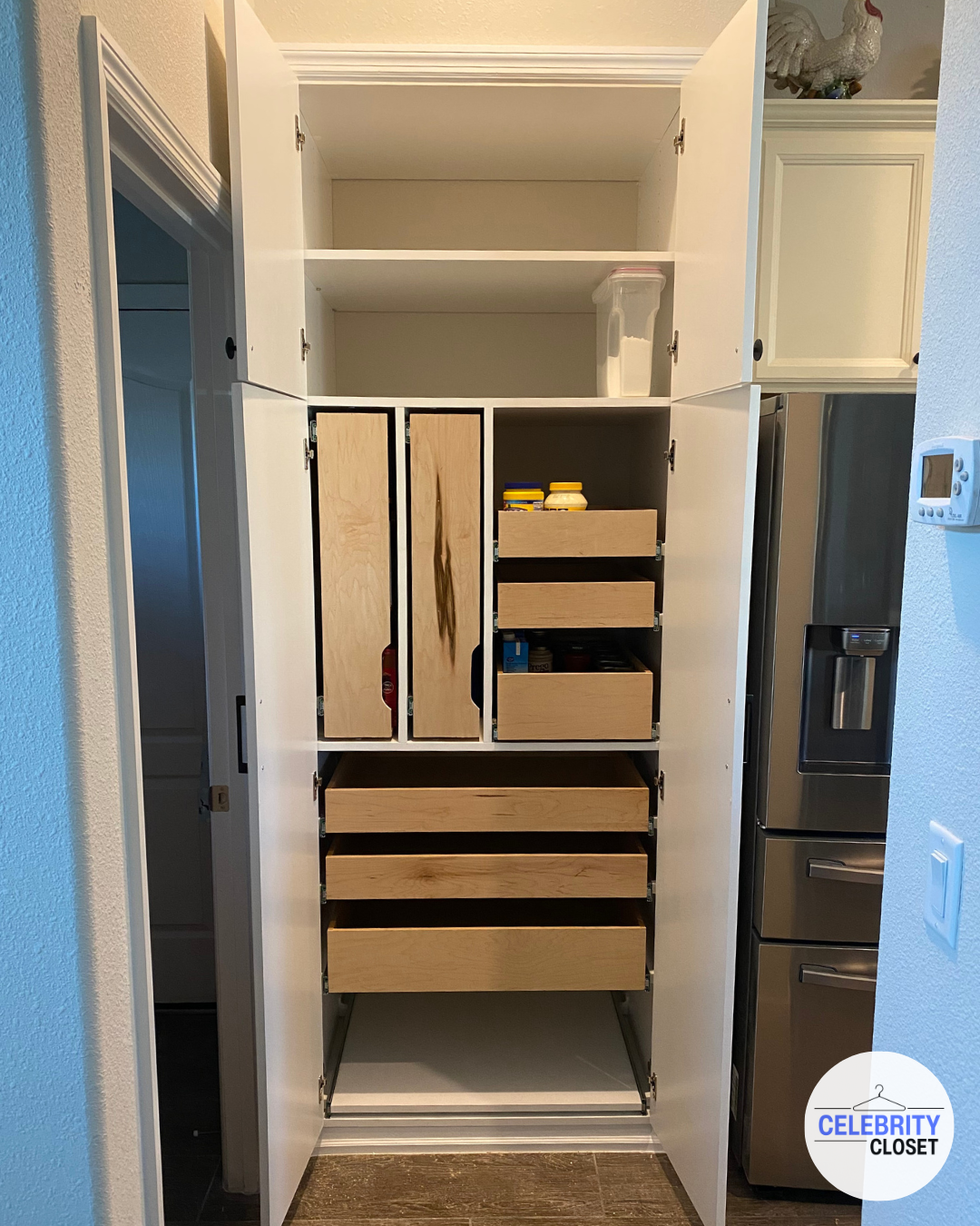 Open kitchen pantry closet with wooden shelves and drawers, some containing food items, next to a stainless steel refrigerator.