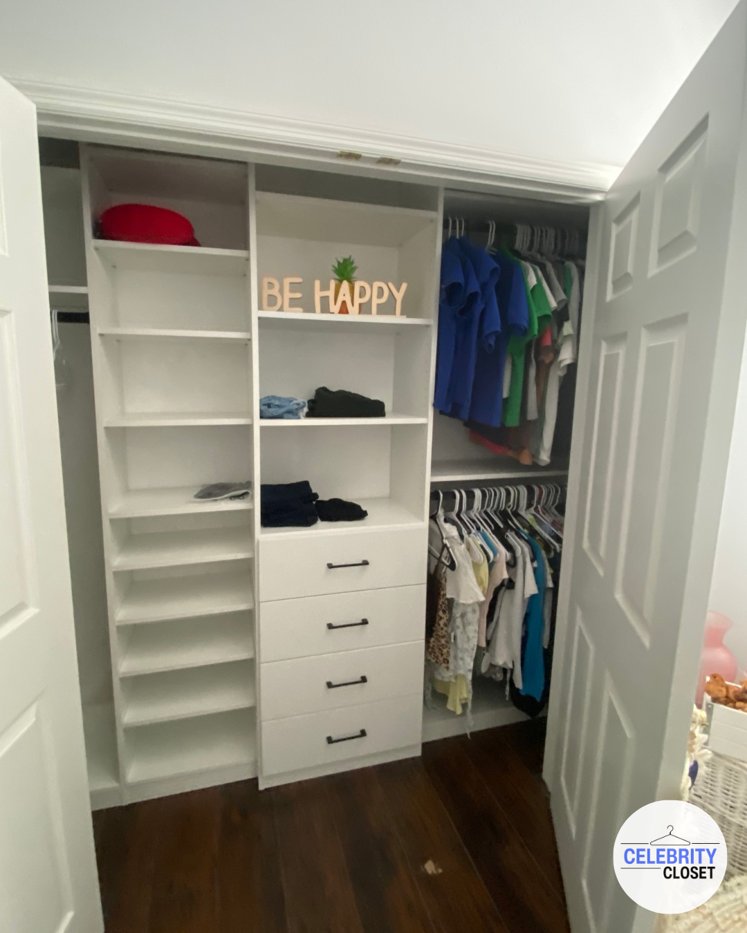 Open closet with shelves and drawers, displaying folded clothes, hanging shirts and pants, with a decorative sign 'BE HAPPY' and a small pineapple decor on the top shelf.