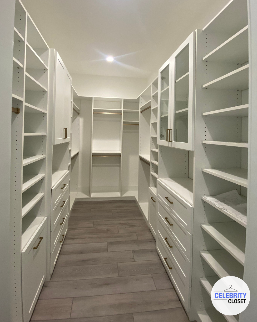 Empty walk-in closet with white built-in shelves and drawers, and wood-look flooring.