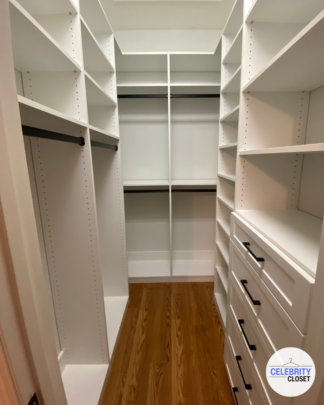 Empty walk-in closet with white shelving and drawers, hardwood floor, and hanging rods.