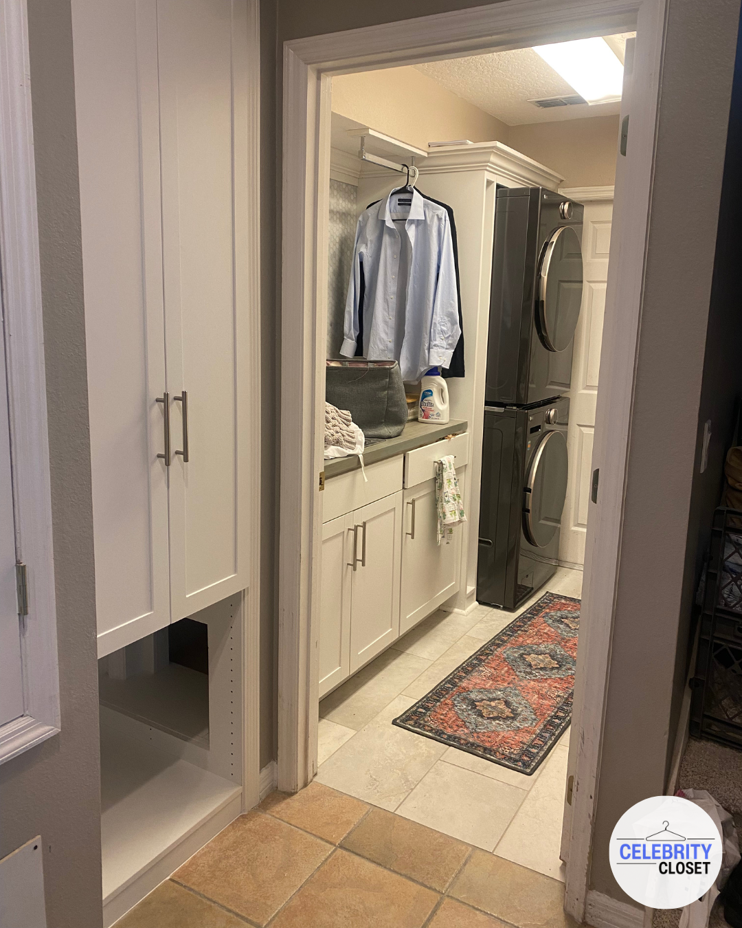 Inside a laundry room with a stackable washer and dryer, white cabinets, a countertop holding a gray bag, laundry detergent, and folded clothes, with a blue dress shirt hanging on a hook, and a small colorful rug on the floor.