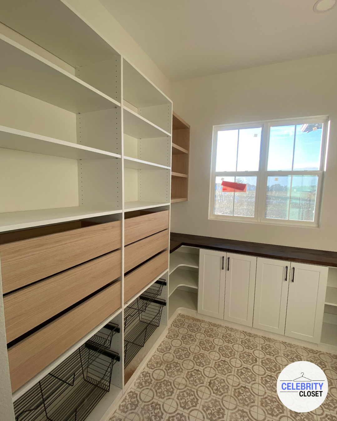Empty storage room with white and wooden shelves, cabinets, and wire baskets. Light-filled window on the right, patterned tile floor, and a dark wooden countertop.