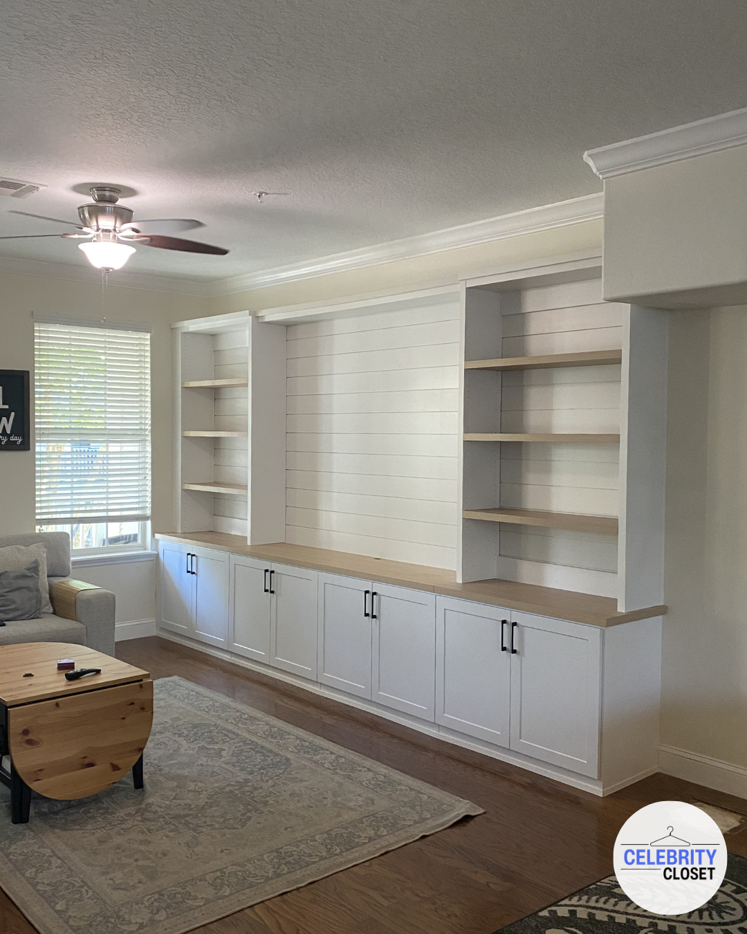 Living room with built-in white shelves and cabinets, a ceiling fan, a window with blinds, and a light-colored sofa with pillows.