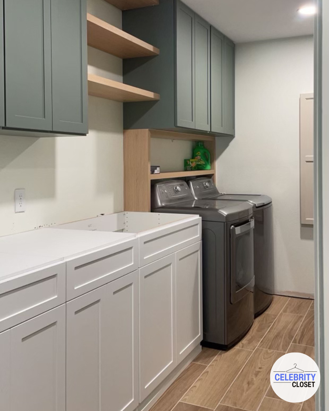 Laundry room with white lower cabinets, gray upper cabinets, open wooden shelves, and a matching wooden back panel. Contains a white utility sink, a front-loading washing machine, a dryer, and a small green laundry detergent bottle.