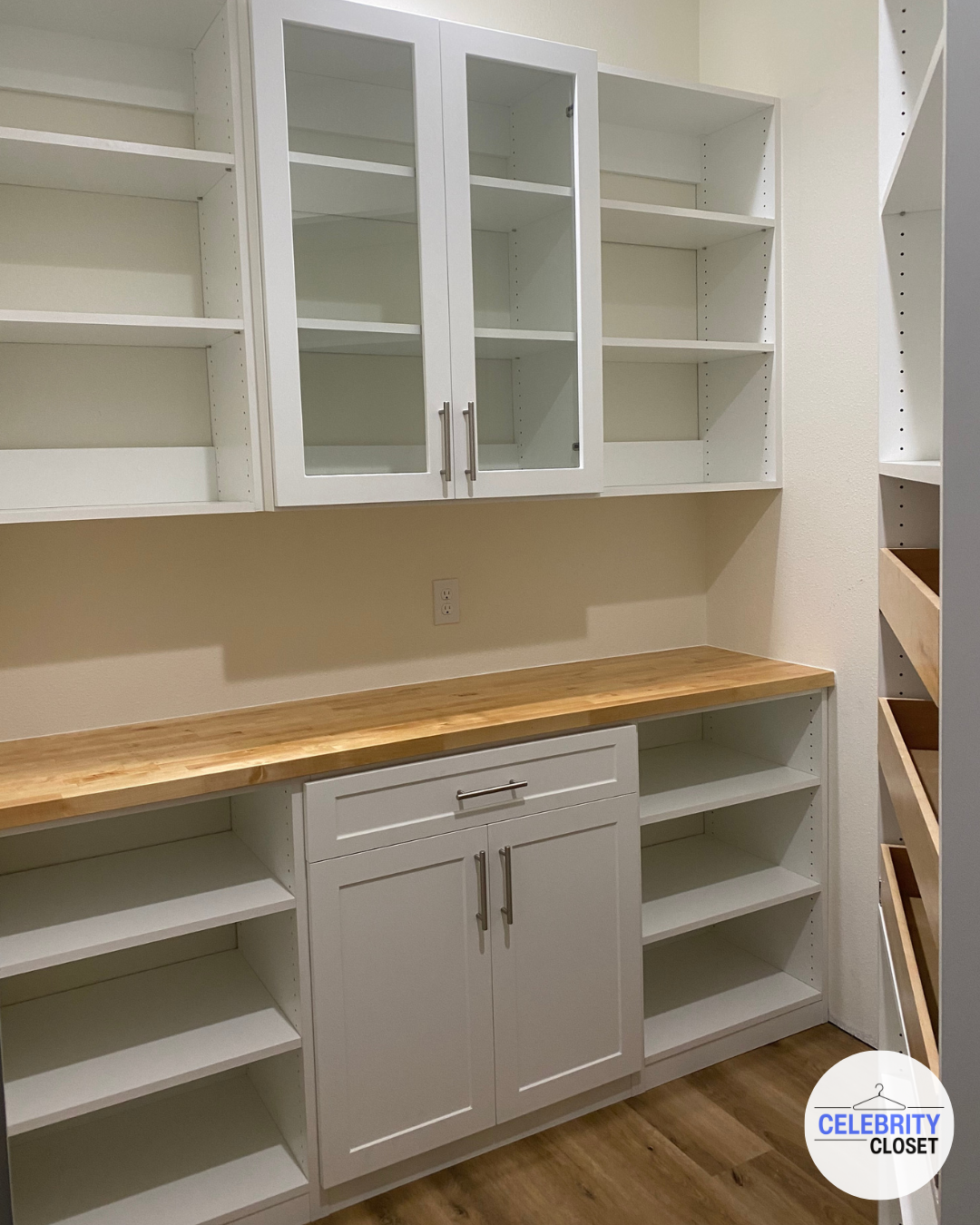 Empty white kitchen cabinets with glass doors and a wooden countertop in a closet or pantry.