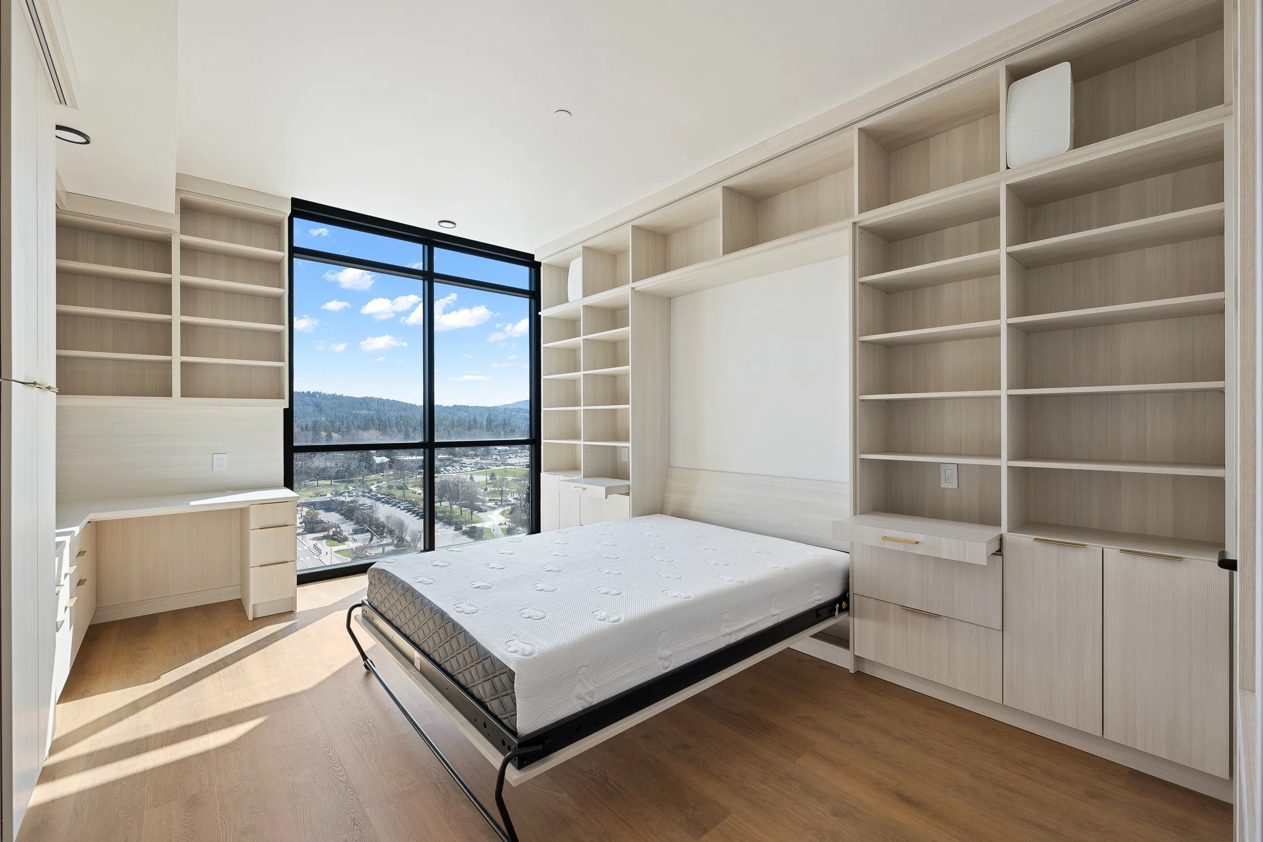 An empty modern bedroom with floor-to-ceiling windows showing a view of a blue sky, white clouds, and distant mountains. The room has built-in light wood shelves and drawers, with a mattress on a metal frame, and natural wood flooring.