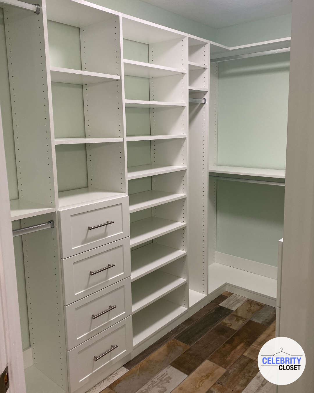 Empty walk-in closet with white built-in shelves, drawers, and hanging rods on mint green walls, with multicolored wood flooring.