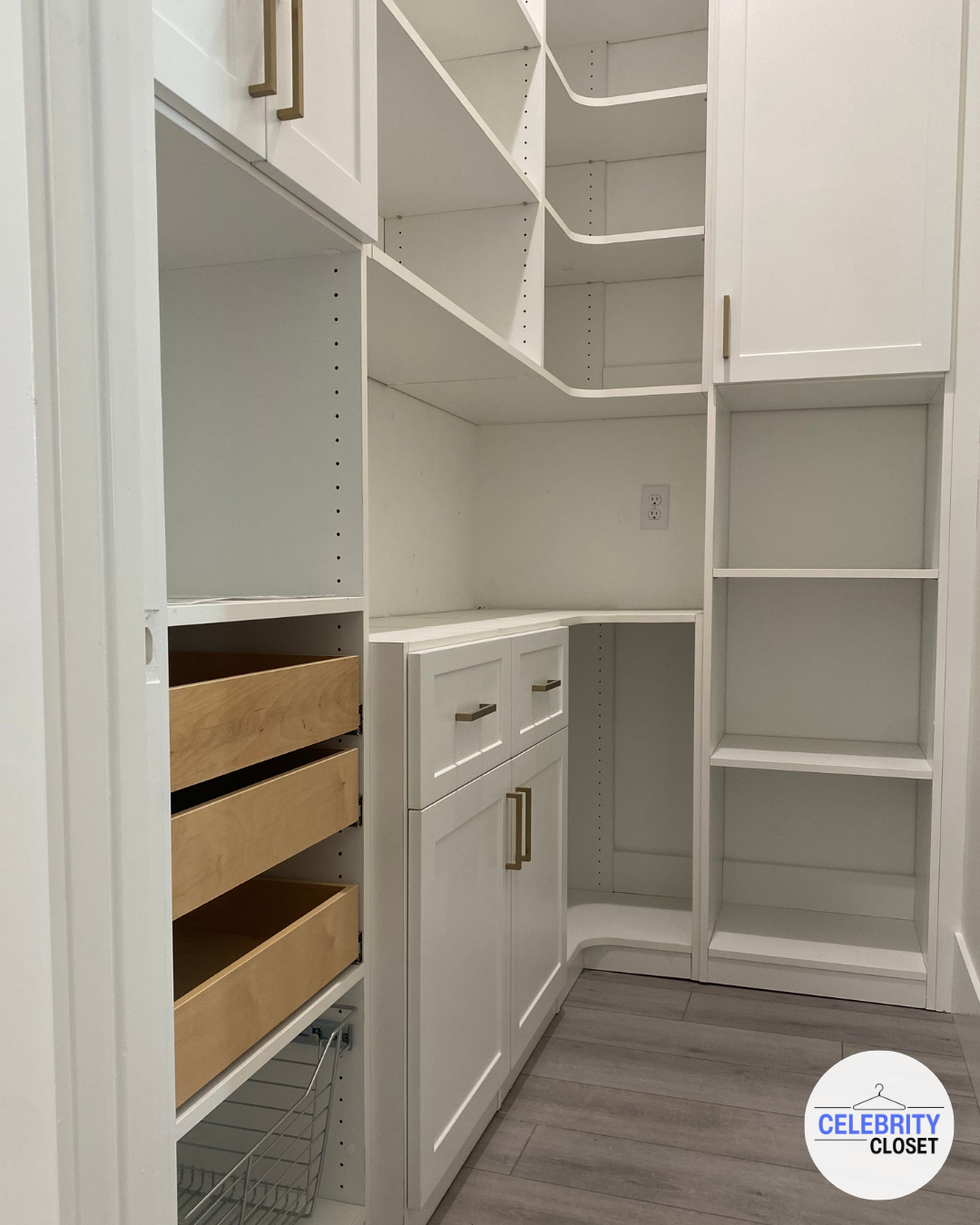 Empty white closet with multiple shelves, drawers, and cabinets, with wood and gold handles, and a three-tier metal basket drawer at the bottom left.