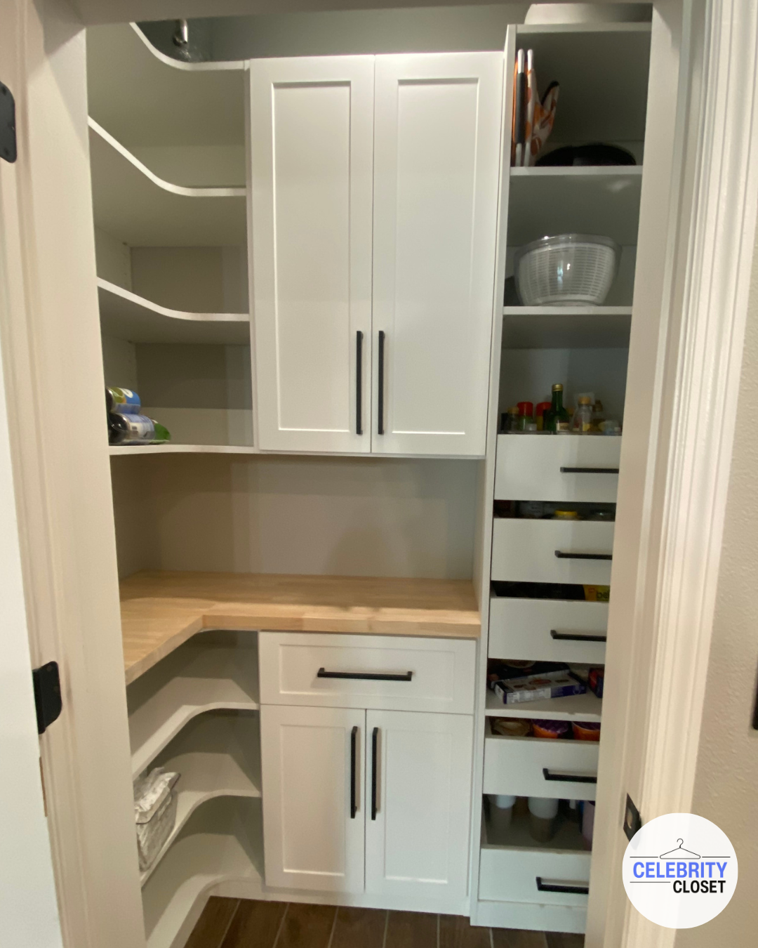 Pantry closet in a home with white shelves, cabinets, and drawers, containing food, snacks, spices, and kitchen items, with a wood countertop.
