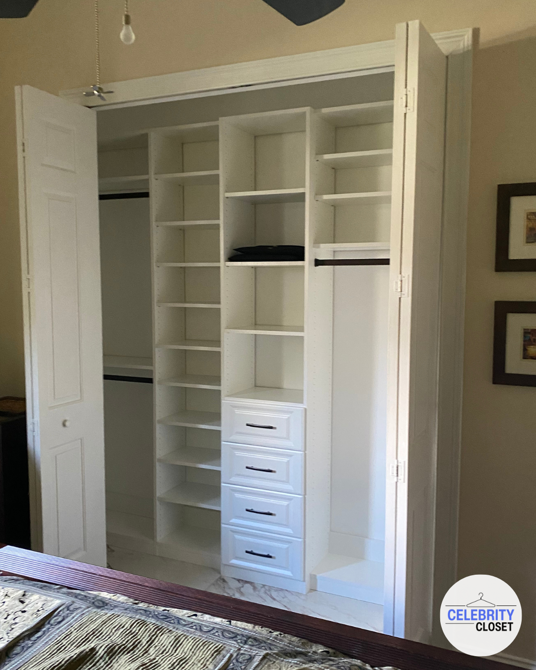 Empty white closet with shelves, drawers, and hanging space, open doors, in a bedroom with a bed and framed pictures on the wall.