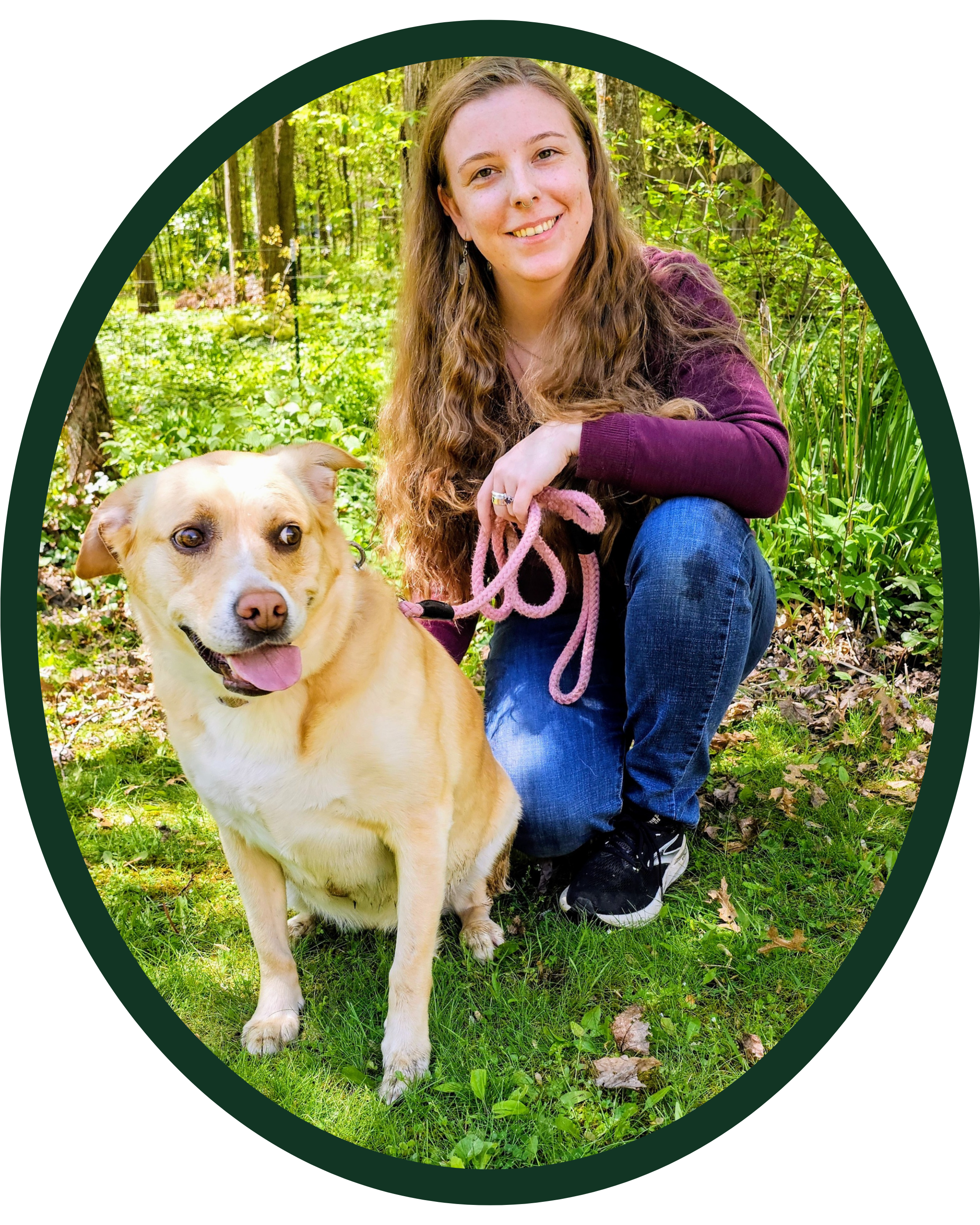A woman with long curly hair, wearing a maroon shirt and blue jeans, kneeling next to a yellow Labrador Retriever dog in a lush green forest.