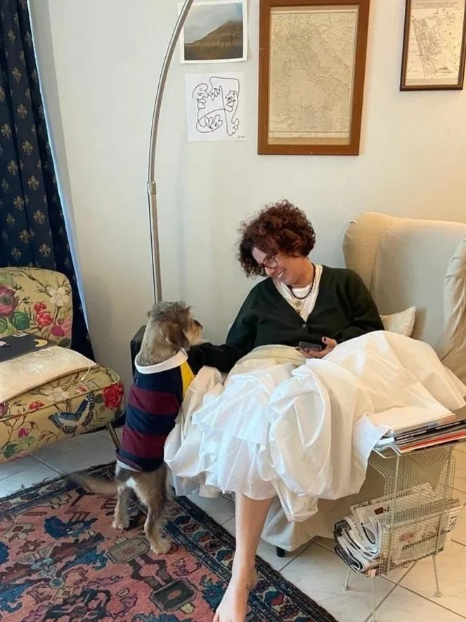 A woman sitting in a chair with a white blanket, looking at her phone, while a small dog in a striped sweater stands on its hind legs, resting its front paws on her lap, indoors in a living room.