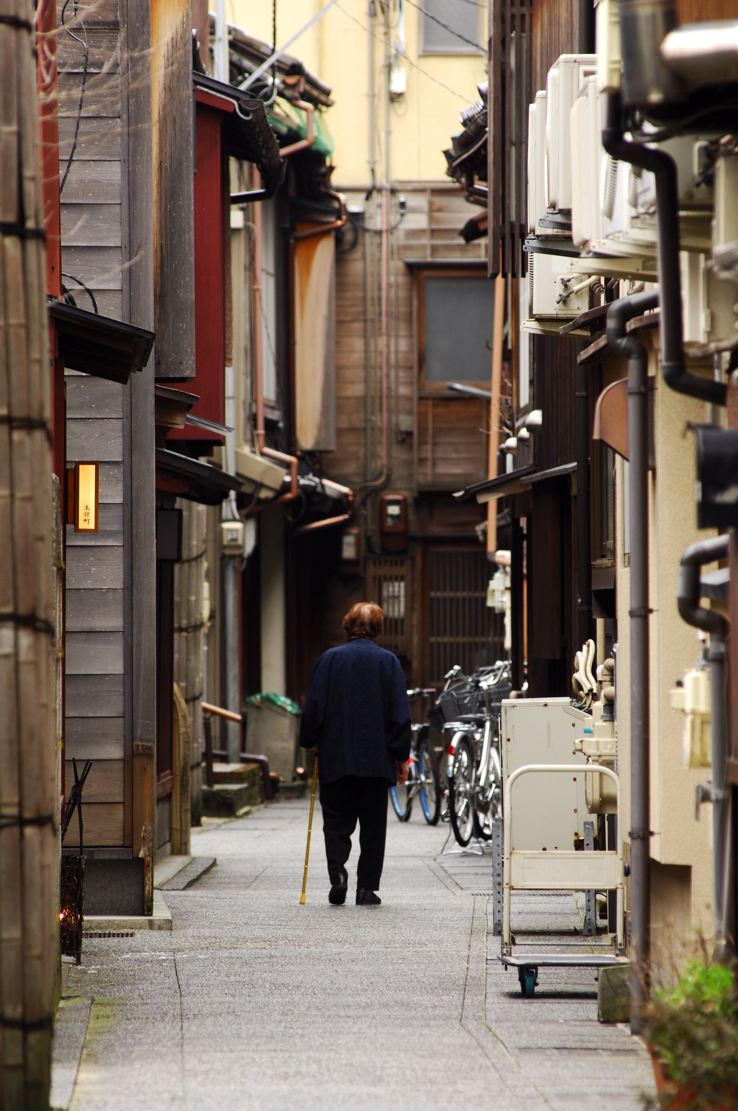 A person walking with a cane down a narrow back street of old wooden houses in Kanazawa, Japan.