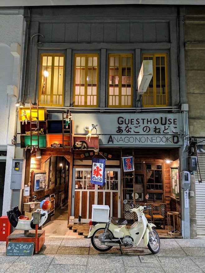Guesthouse Anago no Nedoko lit up at dusk along the Onomichi shōtengai, a vintage scooter parked at its wooden storefront, Japan.