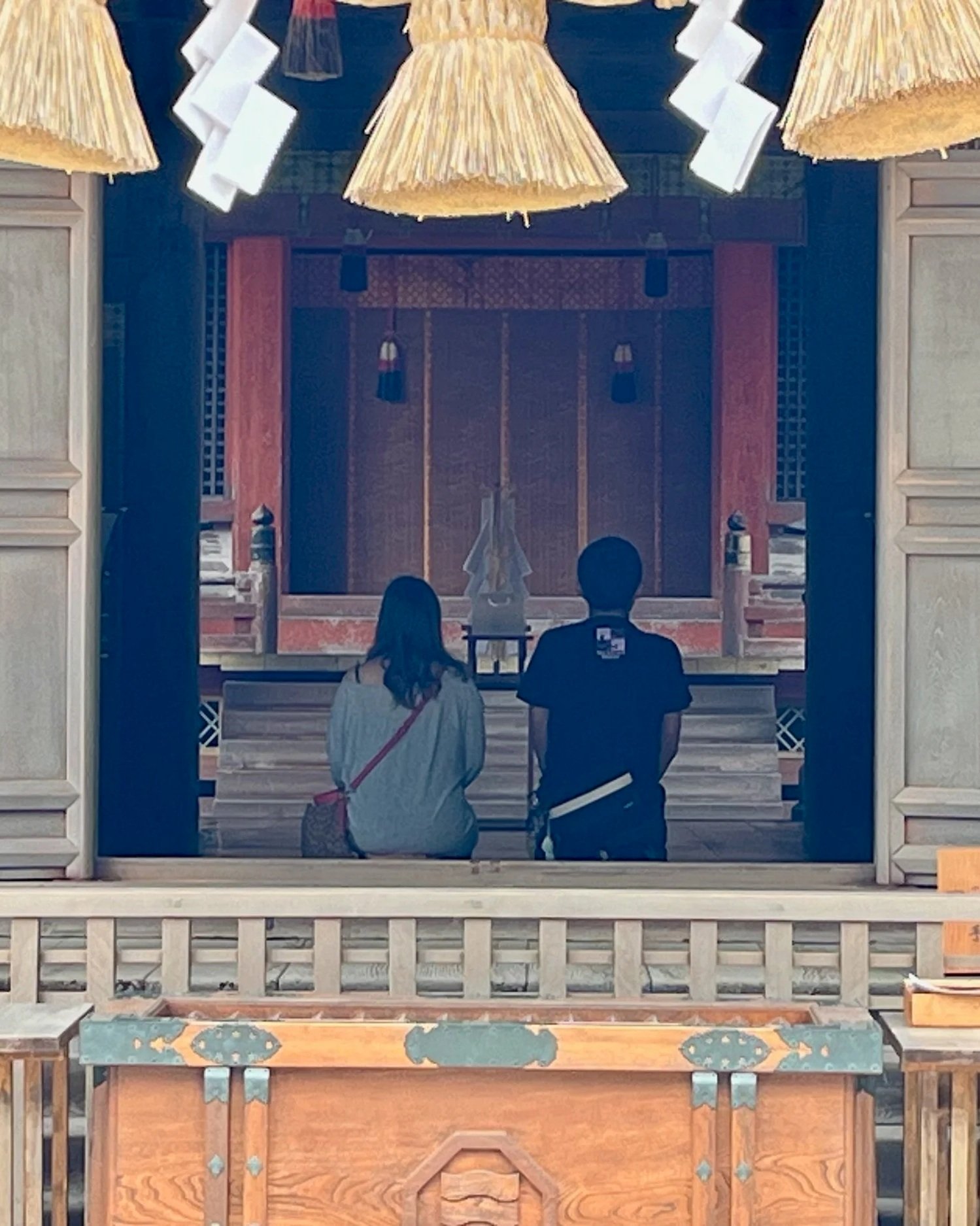 Two people seated inside the hall of Ōyamazumi Shrine on Ōmishima island, Ehime, Japan, framed by a sacred straw rope and paper streamers.