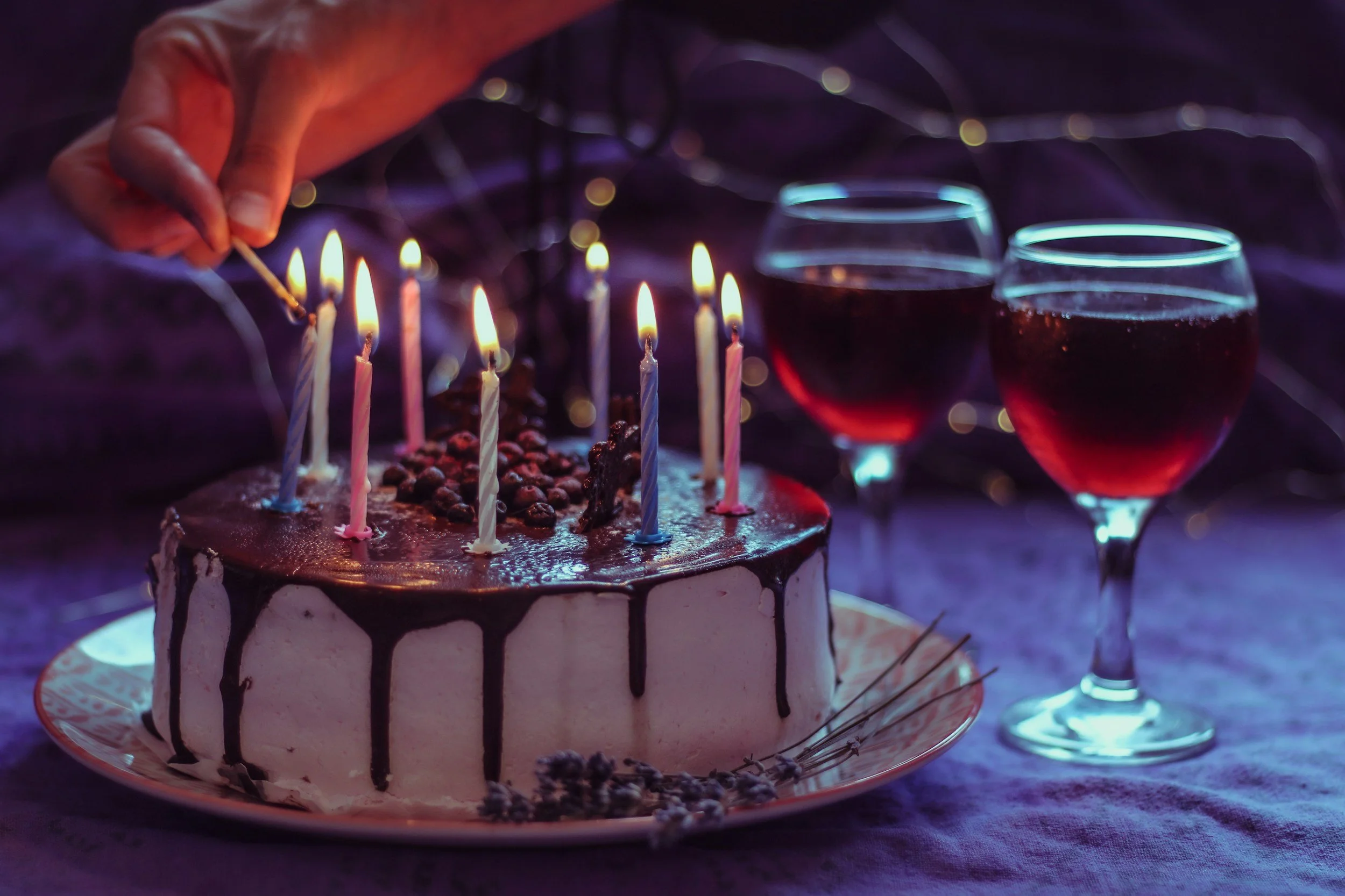 A birthday cake with candles being lit, topped with chocolate and berries, and accompanied by two glasses of red wine on a dark, festive table.