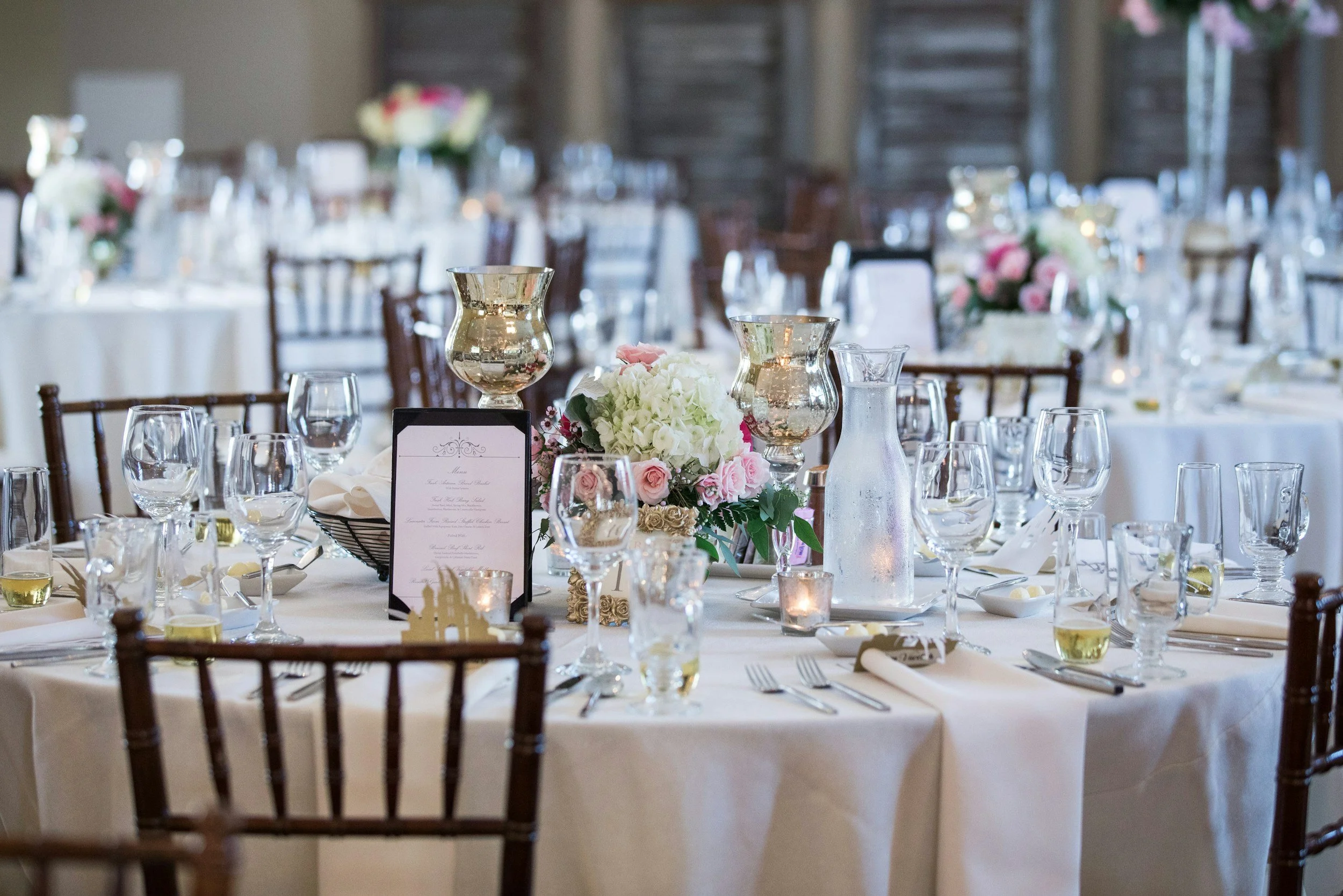 A decorated banquet table set for a formal event, featuring floral centerpieces with pink and white flowers, glass candle holders, wine glasses, cutlery, and a menu card, with additional tables and chairs in the background.