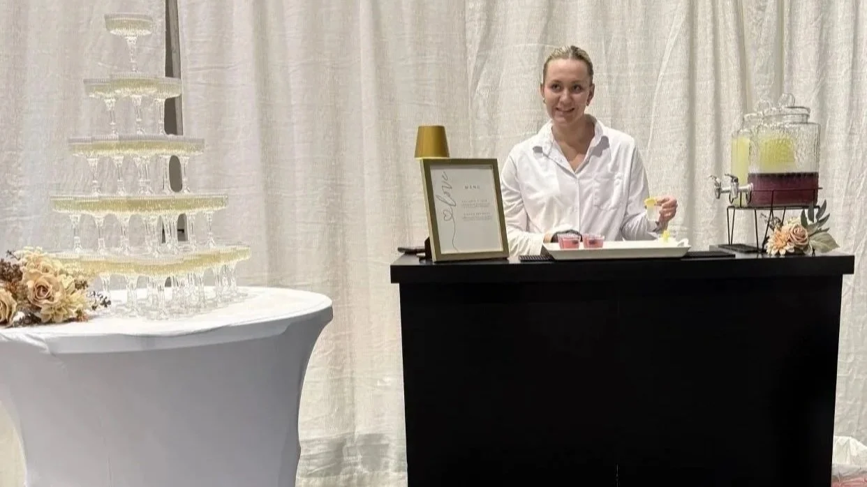 A woman in a white shirt standing behind a black bar counter, with a beverage dispenser and framed sign on the counter, and a white curtain background.