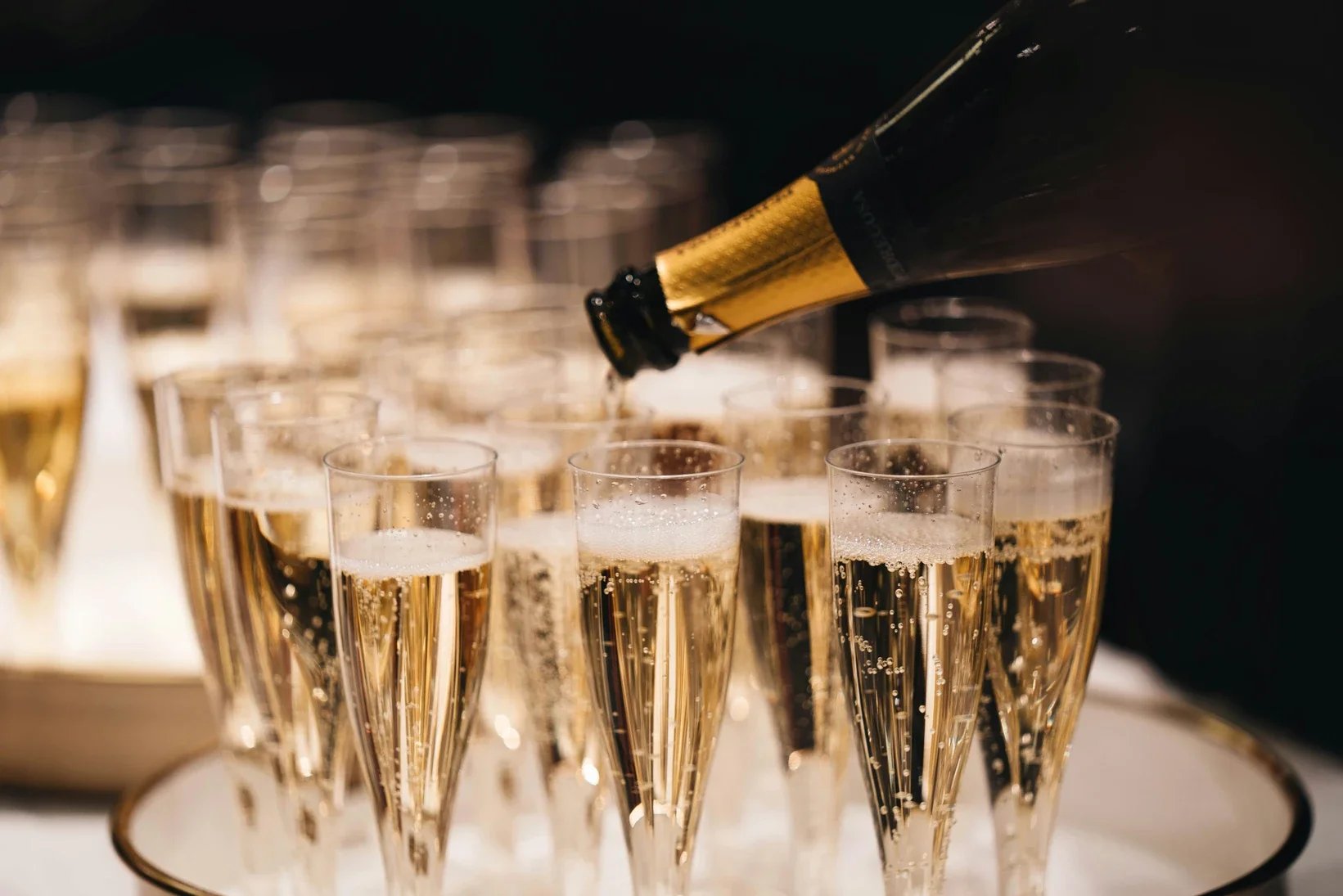 A hand pouring champagne from a bottle into a row of champagne flutes at a celebration or party.