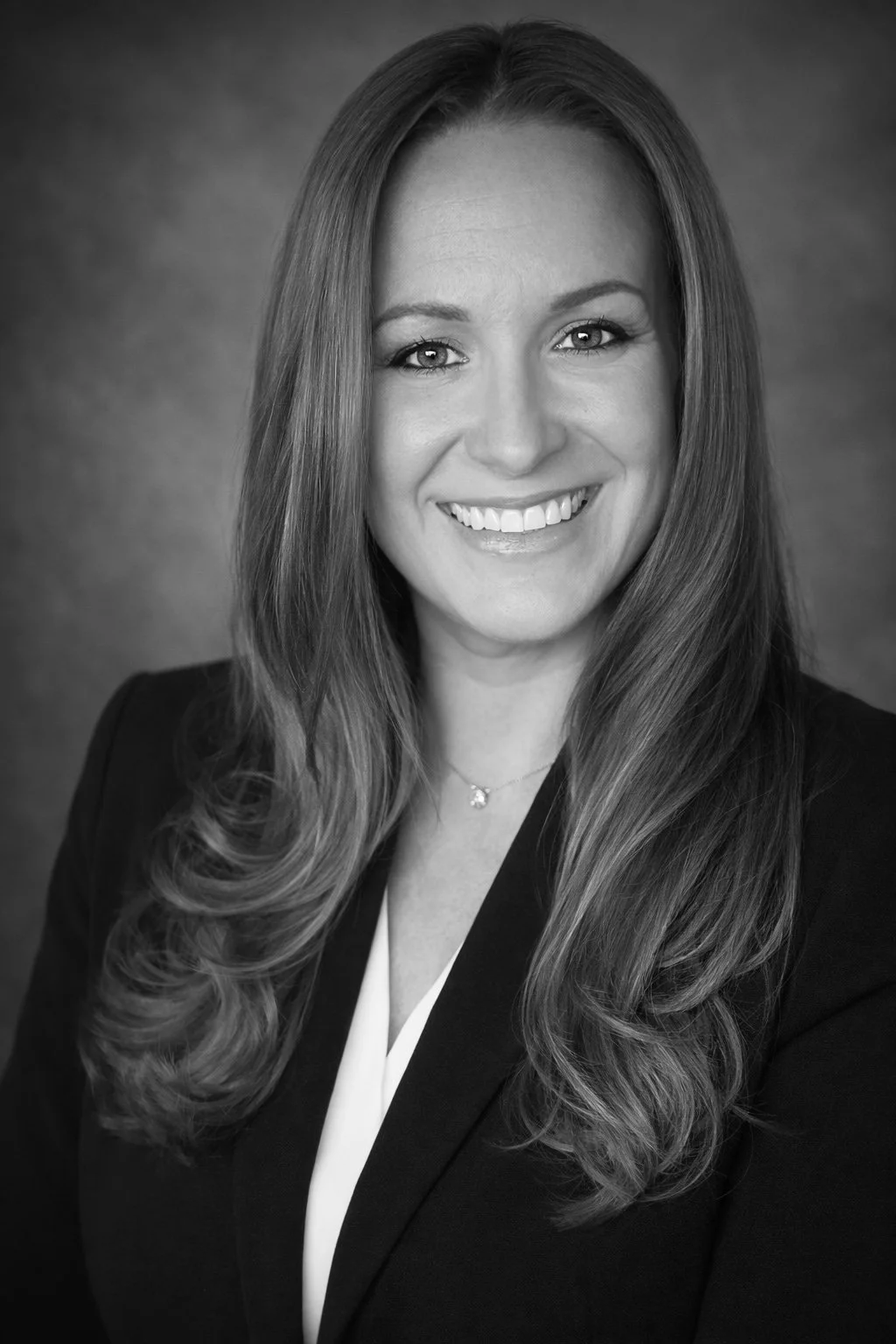 Black and white portrait of a smiling woman with long, wavy hair, wearing a blazer and a necklace.
