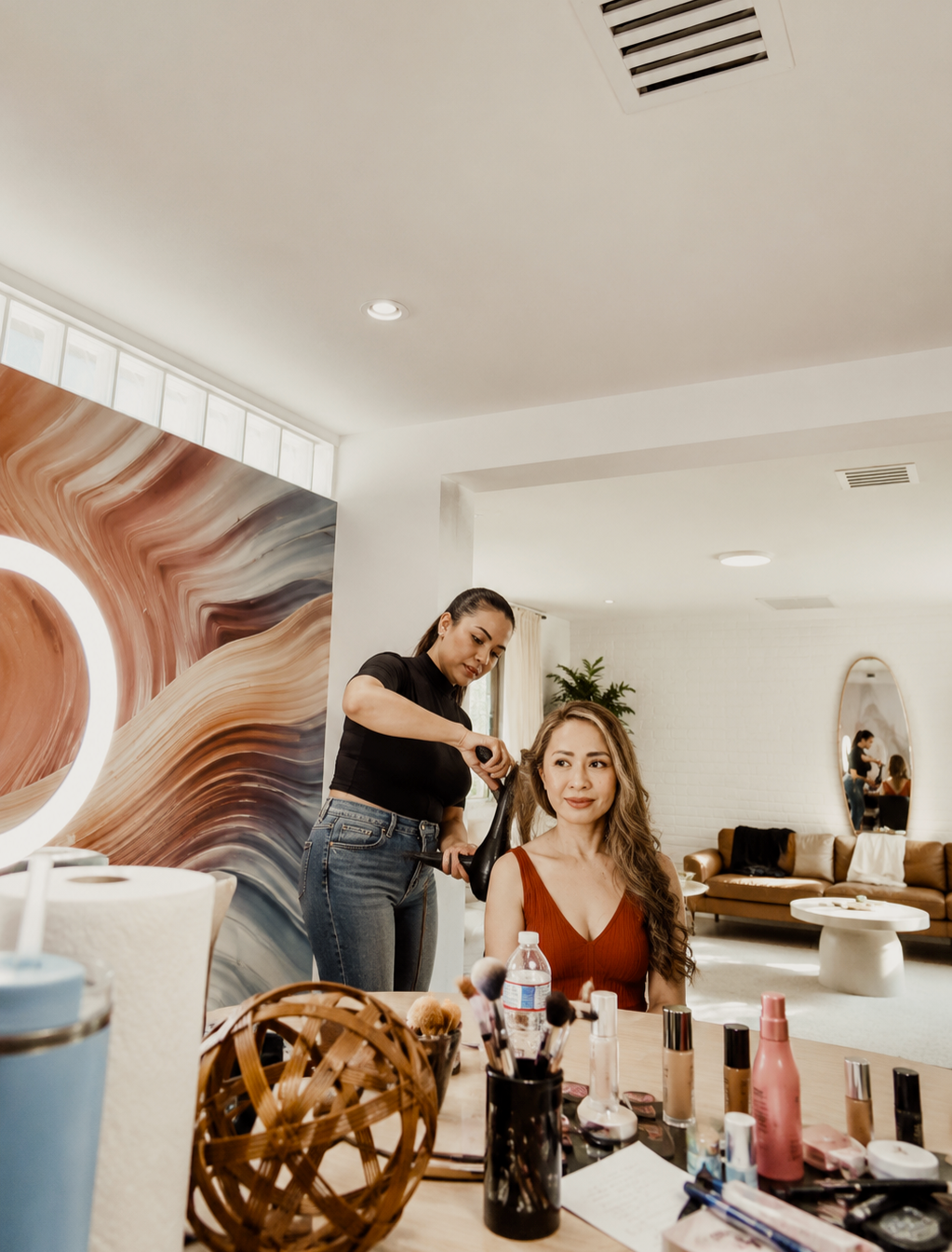 A woman with long wavy hair sitting at a vanity table with makeup products, while a hairstylist blow-dries her hair in a modern, well-lit living room.