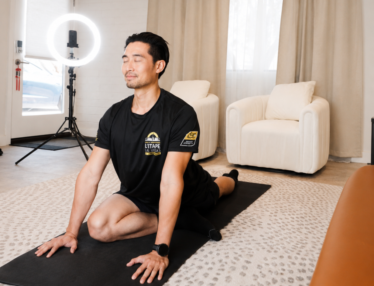A man in black athletic shirt practicing yoga on a black mat inside a bright room with beige carpet and white furniture, with a ring light and camera setup in the background.