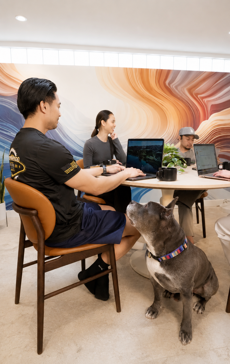 People working on laptops around a table in a modern office with a colorful marble wall mural, and a large dog sitting beside the man in the foreground.