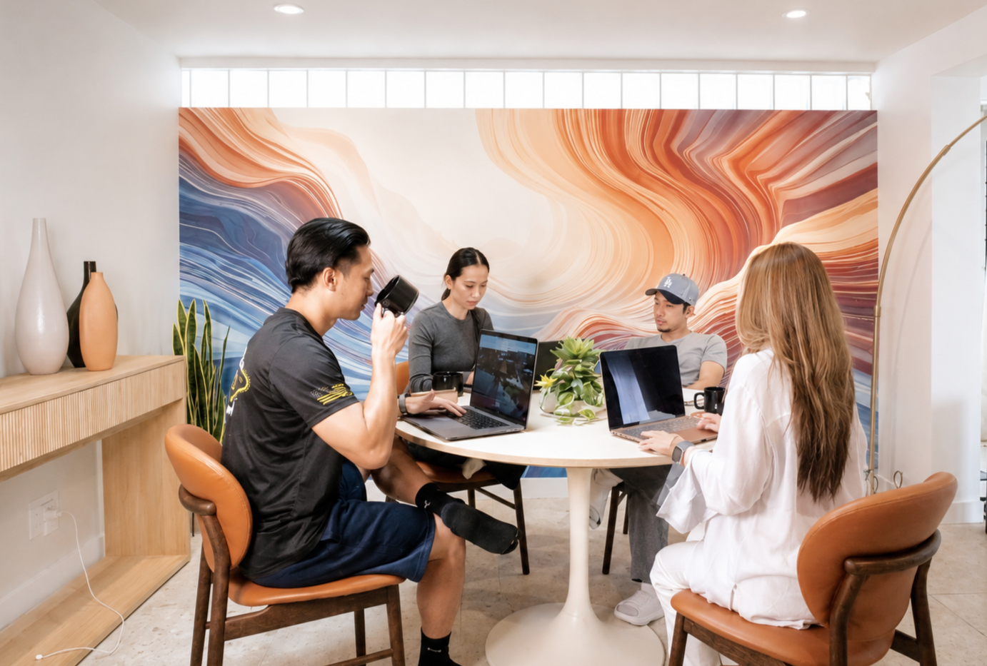 Group of five young adults working and drinking coffee at a round table in a modern, brightly lit space with abstract wall art.