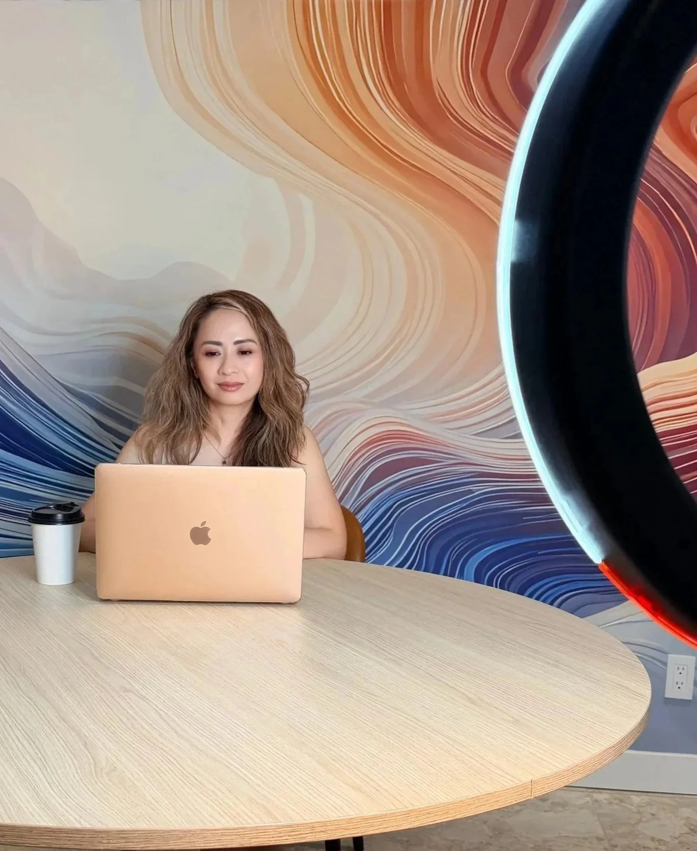 A woman with long wavy hair sitting at a table with a pink Apple MacBook in front of her, a coffee cup to her left, in front of a colorful swirling mural background, with a large ring light partially visible on the right side of the image.