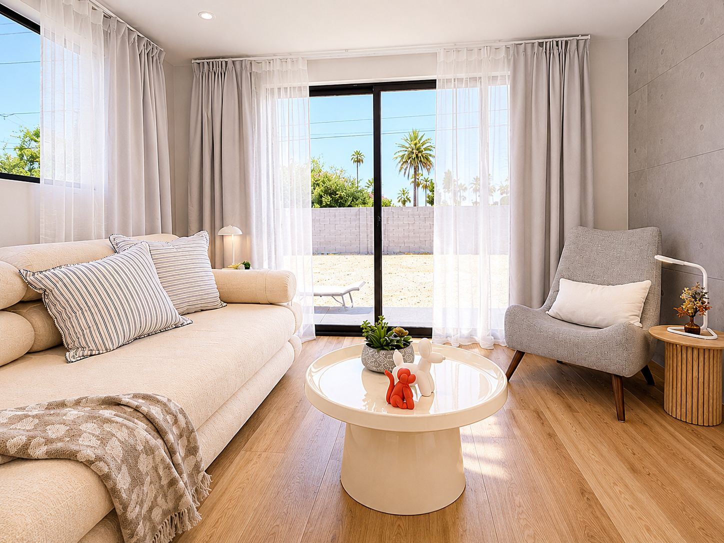 Bright living room with cream sofa, striped pillows, gray armchair with white pillow, round white coffee table with decorative sculptures and plant, side table with flower vase, large sliding glass door with white curtains, and a view of palm trees o